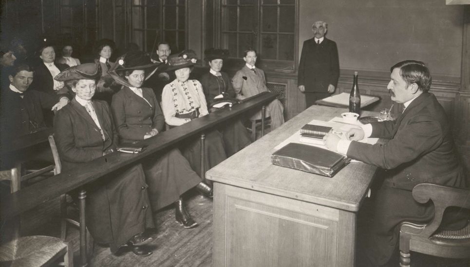 Photo noir et blanc d'une salle de cours, à droite le professeur derrière son bureau, au fond un homme debout, à gauche au premier rang, 4 femmes et un homme, au deuxième rang, 5 autres personnes.
