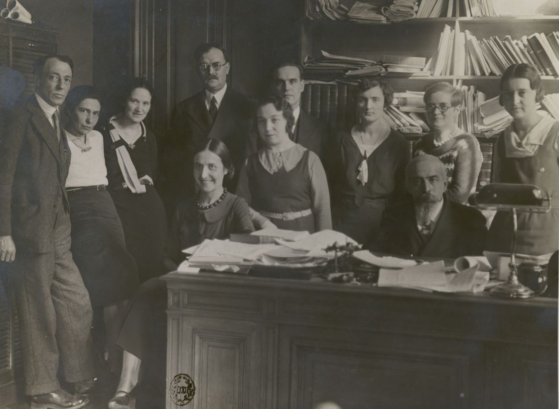 Photo en noir et blanc de 11 personnes travaillant à la bibliothèque de la Sorbonne en tenue des années 1930 posant dans un bureau avec des étagères remplies de papiers et de livres.